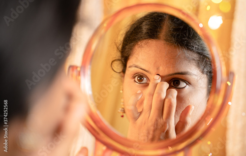 Indian married Woman applying Bindi, sindoor or decorative mark to forehead in front of mirror during festival celebrations with decoration lights as background. photo