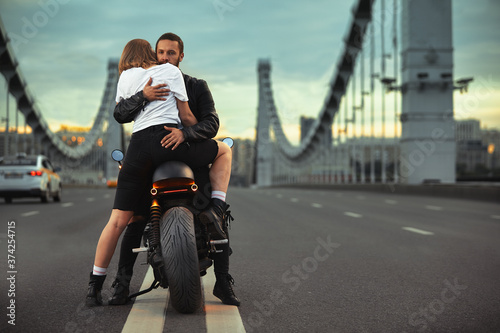 Young sexy woman hugging cute man in stylish black leather jacket, sitting on sports motorcycle on the bridge in the city on sunset and kissing