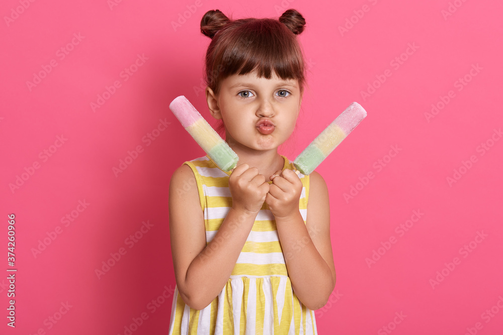 Girl with two water ice cream looking at camera and making grimace, pout lips, wearing striped summer dress, standing isolated over pink background. Girl with two water ice cream looking at camera and making grimace, pout lips, wearing striped summer dress, standing isolated over pink background.