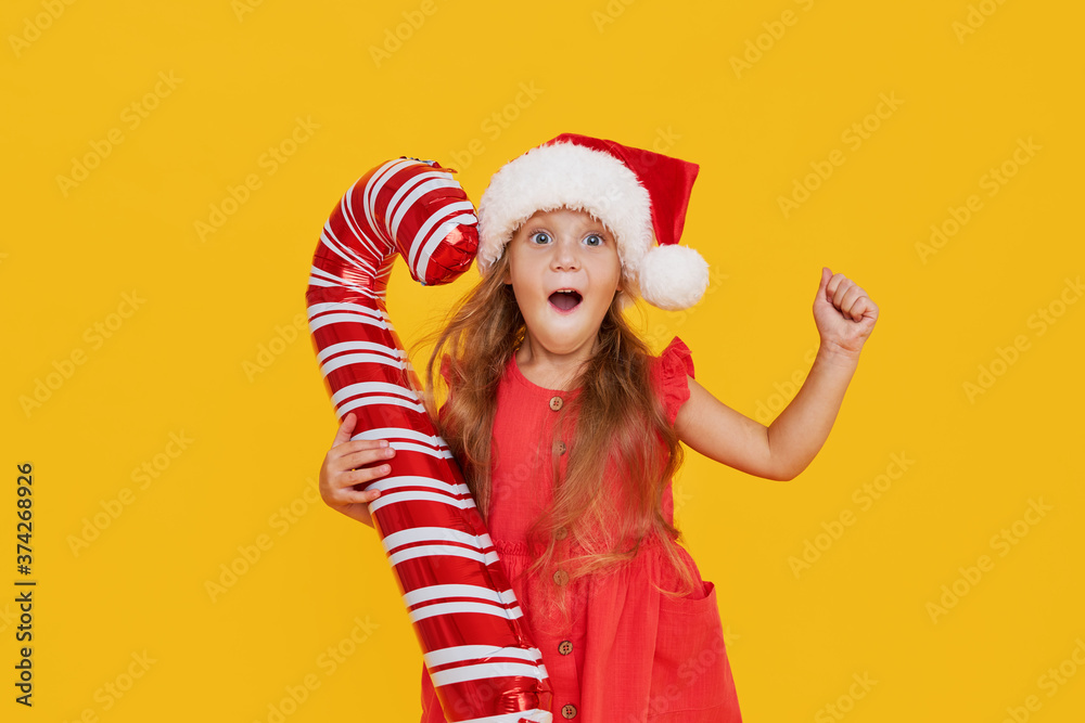 A cute little girl child in a fancy dress and a Santa hat holds an inflatable shape of a candy cane on a yellow background. 2021 new year holidays concept. points to the right A cute little girl child in a fancy dress and a Santa hat holds an inflatable shape of a candy cane on a yellow background. 2021 new year holidays concept. points to the right