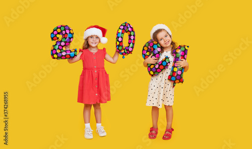 Cheerful children in Santa hats holding colorfull numbers 2021 on yellow background. Happy New Year party with balloons.