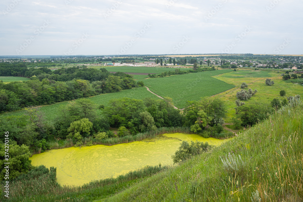Wonderful rural landscape. Valley view with green lake, fields and village on blue sky background