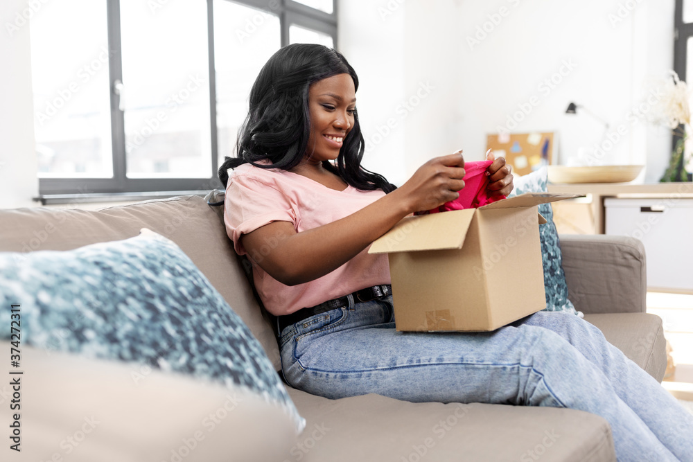 delivery, shipping and people concept - happy young african american woman opening parcel box with clothes at home