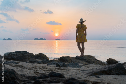 Young caucasian woman in hat and yellow dress on the sandy beach enjoying the sunset