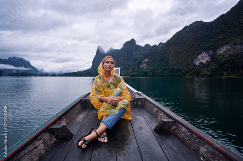 Travel by Thailand. Young woman in raincoat sailing Khao Sok National Park Cheow Lan lake on traditional longtail boat.