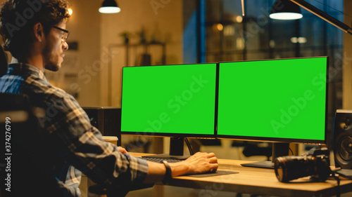 Over the Shoulder: Creative Designer Sitting at His Desk Uses Desktop Computer with Two Green Mock-up Screens. Professional Office Employee Working Late in the Evening in His Studio 