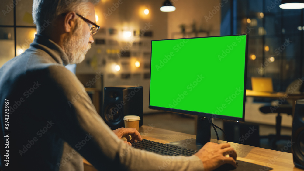 Creative Middle Aged Designer Sitting at His Desk Uses Desktop Computer with Green Mock-up Screen. In the Evening Creative Employe Working on Computers in the Office with Cityscape Window View Creative Middle Aged Designer Sitting at His Desk Uses Desktop Computer with Green Mock-up Screen. In the Evening Creative Employe Working on Computers in the Office with Cityscape Window View