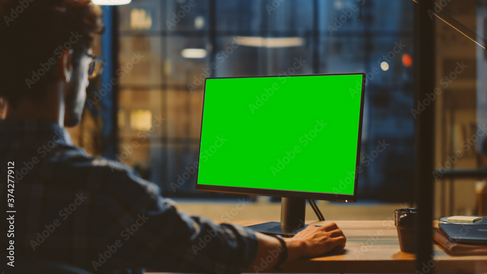 Obraz premium Over the Shoulder: Creative Young Man Sitting at His Desk Using Desktop Computer with Mock-up Green Screen. Evening in the Stylish Office Studio with City Window View
