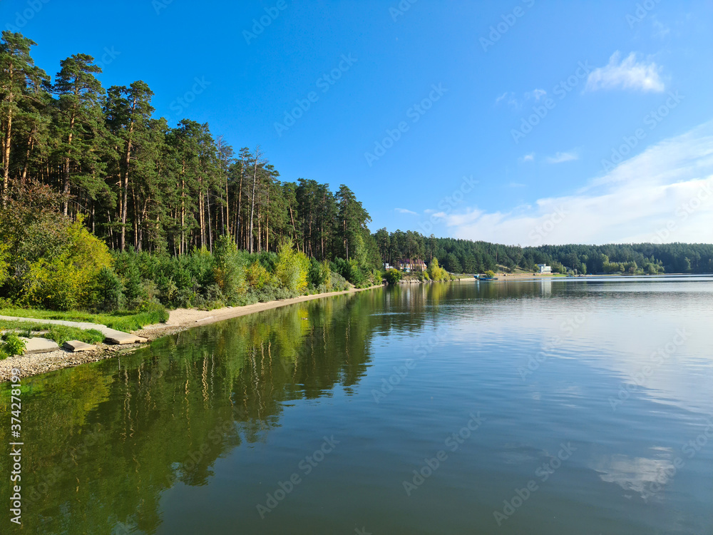 Obraz premium Landscape of the river Bank with bushes, trees and a small beach. Forest along the river with a reflection in the water against the bright blue sky.