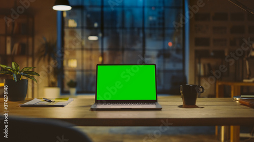 Mock-up Green Screen Laptop Standing on the Desk in the Modern Creative Office. In the Background Warm Evening Lighting and Open Space Studio with City Window View. © Gorodenkoff