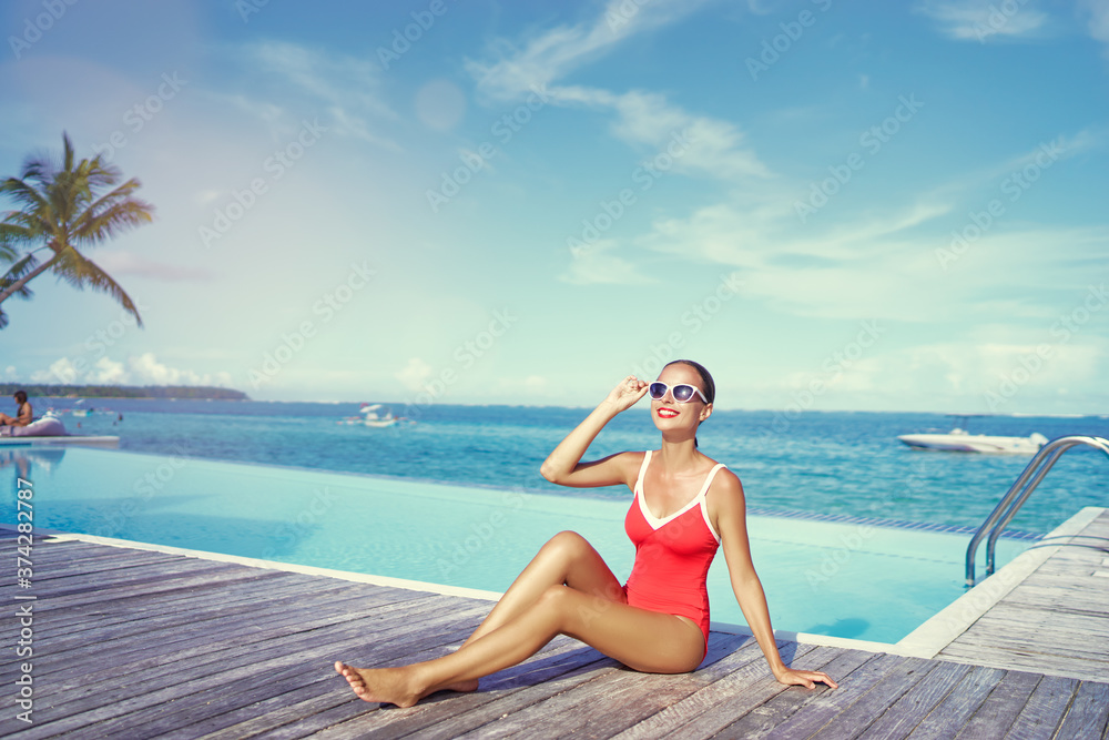 Summer vacation and fashion. Beautiful young woman in red swimsuit near swimming pool on tropical beach.