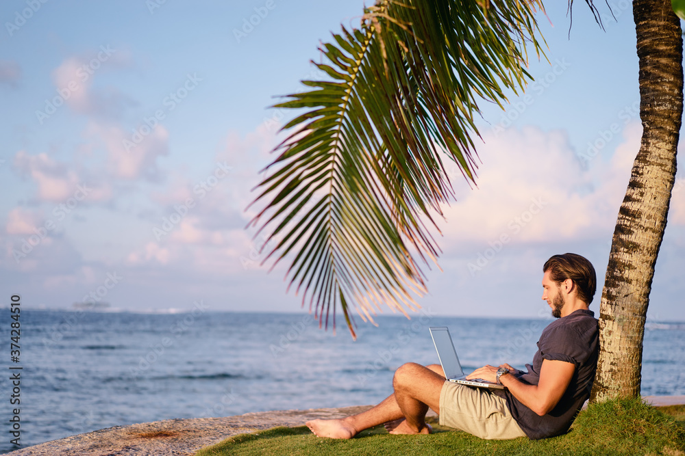 Work and vacation. Young man working on laptop computer on the tropical beach under the palm tree.