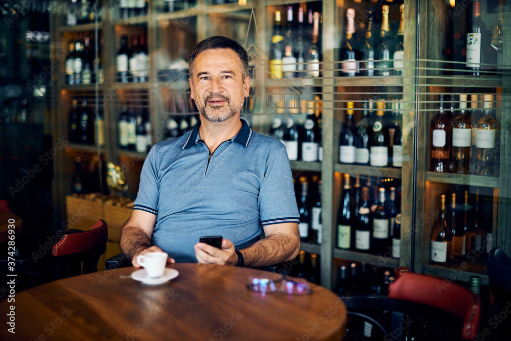 Senior adult sitting in winery, using smart phone on a coffee break while looking at camera.