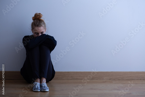 Young woman with anxiety disorder wearing dark clothes sitting on the floor, copy space on empty wall