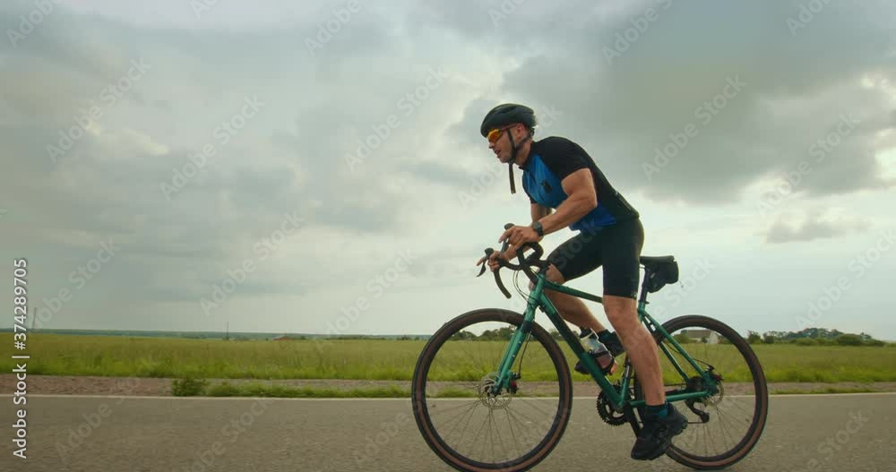 The cyclist is riding along the highway and increasing speed. He's riding uphill. Training on a bicycle. Fields in the background. 4K The cyclist is riding along the highway and increasing speed. He's riding uphill. Training on a bicycle. Fields in the background. 4K