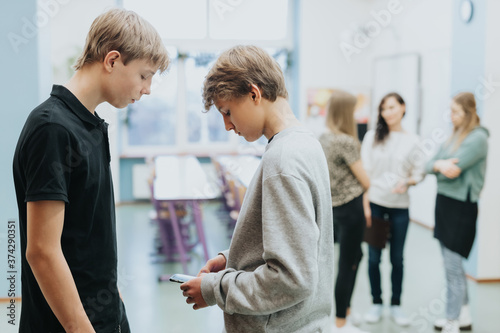Two teenagers are standing together at the side of the classroom, one of them is checking something on a mobile phone