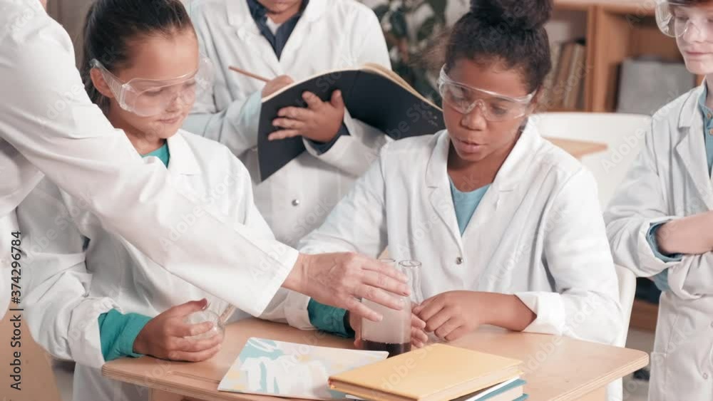 Slowmo tilt up shot of female chemistry teacher doing experiment with curious schoolchildren in lab coats in modern classroom Slowmo tilt up shot of female chemistry teacher doing experiment with curious schoolchildren in lab coats in modern classroom