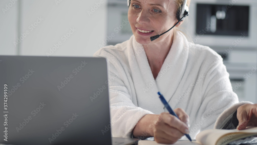 Mature woman with headset and laptop having video conference and taking notes at home office Mature woman with headset and laptop having video conference and taking notes at home office
