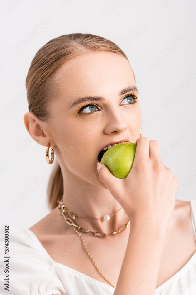 blonde woman biting green apple and looking away isolated on white