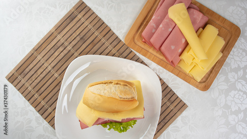 Mortadella sandwich, lettuce and cheese on a white plate on a mat and slices of mortadella and cheese next to it on a cold board on a table with white towel, selective focus. photo