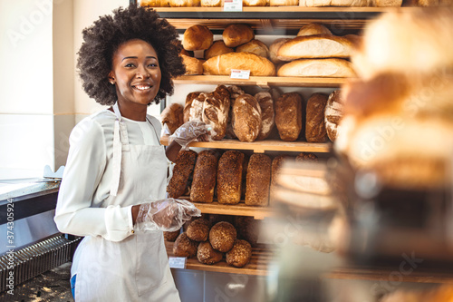 Smiling baker woman standing with fresh bread at bakery. Happy african woman standing in her bake shop and looking at camera.Baker with breads in background. Beautiful and smiling woman at bakery shop