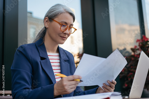 Pensive mature businesswoman reading contract, planning project, brainstorming. Portrait of middle aged asian secretary reading, working with documents, sitting at workplace 