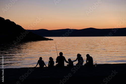 Black silhouettes of people against the background with sunset by the sea. Family is sitting on edge of the pier and fishing. Sveti Juraj, Croatia