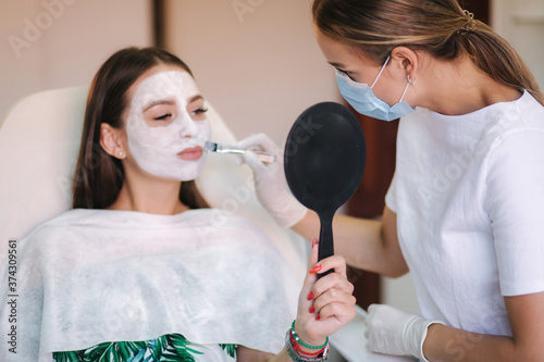 Cosmetologys specialist applying white facial mask using brush. Making skin hydrated and face glowing and skin. Hulf face with clay mask. Attractive young brunette relaxing with closed eyes and photo