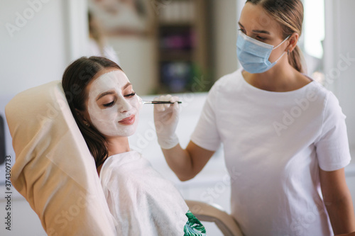 Cosmetologys specialist applying white facial mask using brush. Making skin hydrated and face glowing and skin. Hulf face with clay mask. Attractive young brunette relaxing with closed eyes and photo