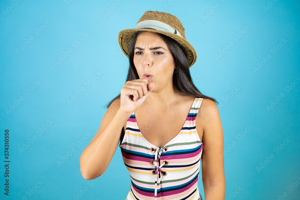 Young beautiful woman wearing swimsuit and hat over isolated blue background with her hand to her mouth because she's coughing Young beautiful woman wearing swimsuit and hat over isolated blue background with her hand to her mouth because she's coughing