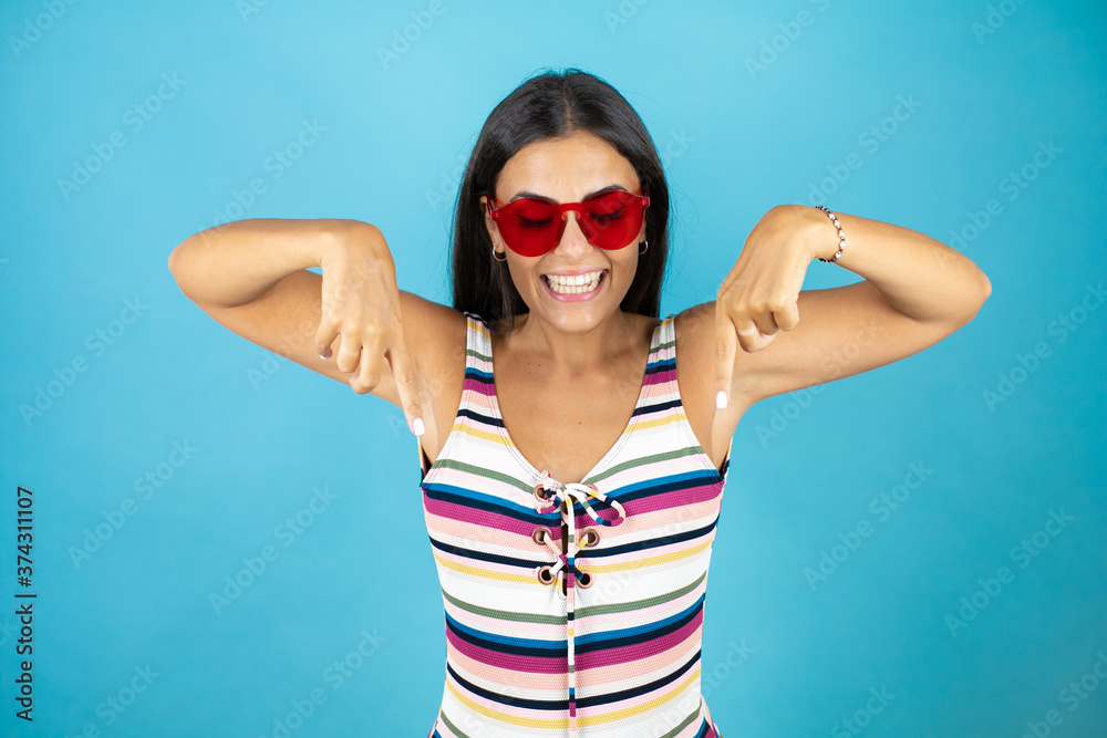Young beautiful woman wearing swimsuit and sunglasses over isolated blue background smiling, looking down and pointing with fingers and raised arms Young beautiful woman wearing swimsuit and sunglasses over isolated blue background smiling, looking down and pointing with fingers and raised arms