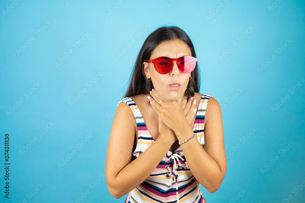 Young beautiful woman wearing swimsuit and sunglasses over isolated blue background with her hand to her mouth because she's coughing Young beautiful woman wearing swimsuit and sunglasses over isolated blue background with her hand to her mouth because she's coughing