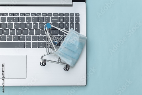 Shopping trolley in medical mask on laptop keyboard. Blue background. Covid-19 pandemic. Online shopping. Top view