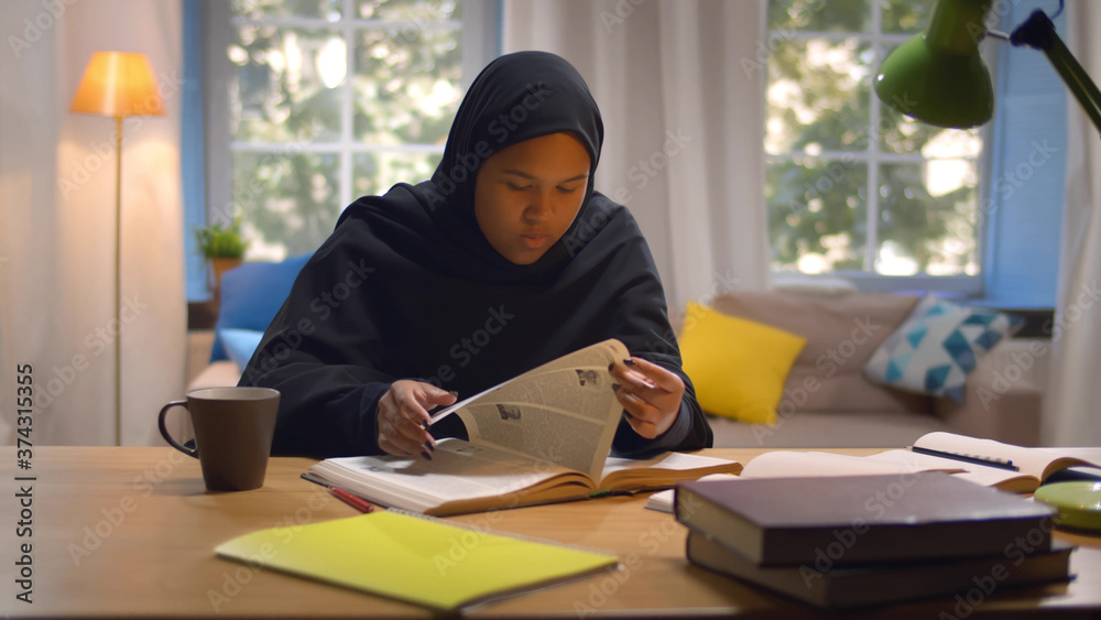 Female muslim college student in hijab sitting at desk and reading book preparing for exam indoors