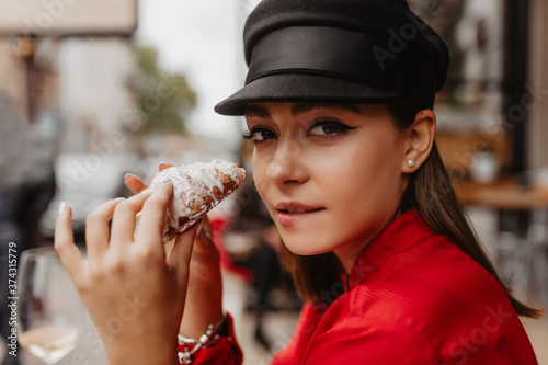 Brunette girl bites her lip at sight of bun with icing. Close-up portrait of stylish blogger traveling in France