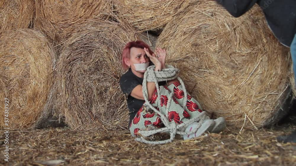 Stock woman kidnapped and raped tied with a rope and gagged with adhesive tape in a bales of hay shed that is intimidated by captor | Adobe Stock