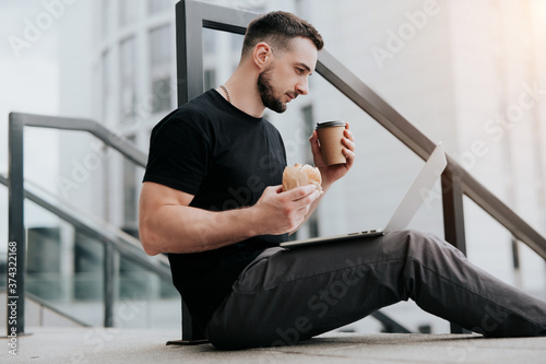 young handsome man sitting on the stairs in the street, works in his laptop while drinking coffee and biting cheeseburger during lunch break, multitasking photo