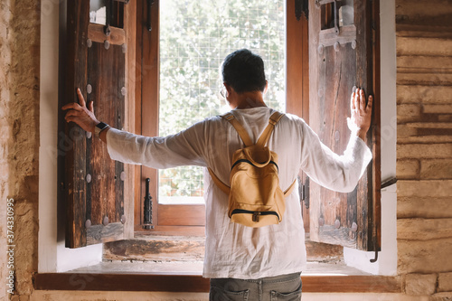 Rear foreground of a man with a backpack who opening two old traditional wooden window shutters to let in the sunlight. Rural and proximity tourism concept photo