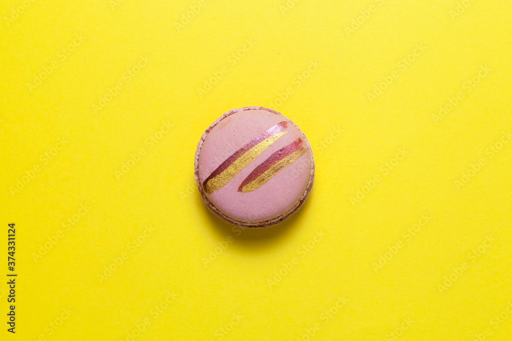 Obraz premium One pink macaron or macaroon with shiny golden stripes lies on a yellow background. Top view, copy space