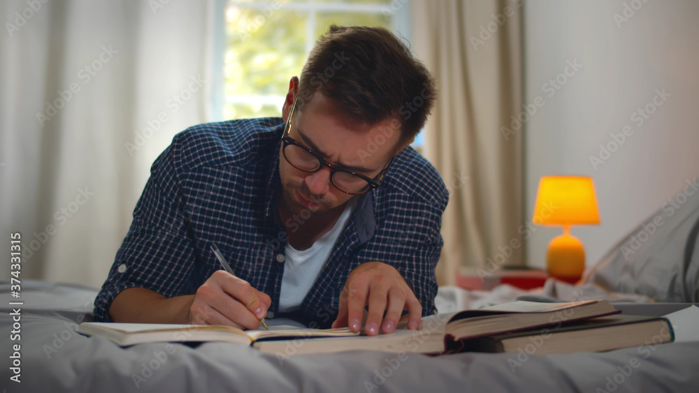 College student lying on bed at home and writing in copybook and reading books when doing homework College student lying on bed at home and writing in copybook and reading books when doing homework