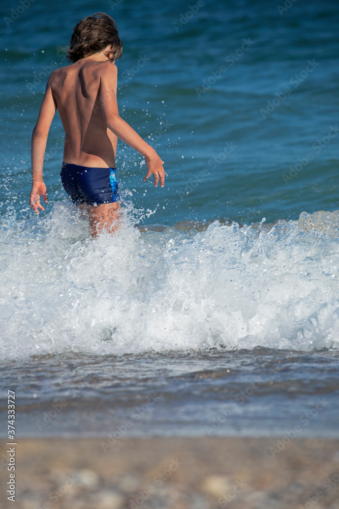 happy boy with his back walks into the sea with splashes and waves, vertical format