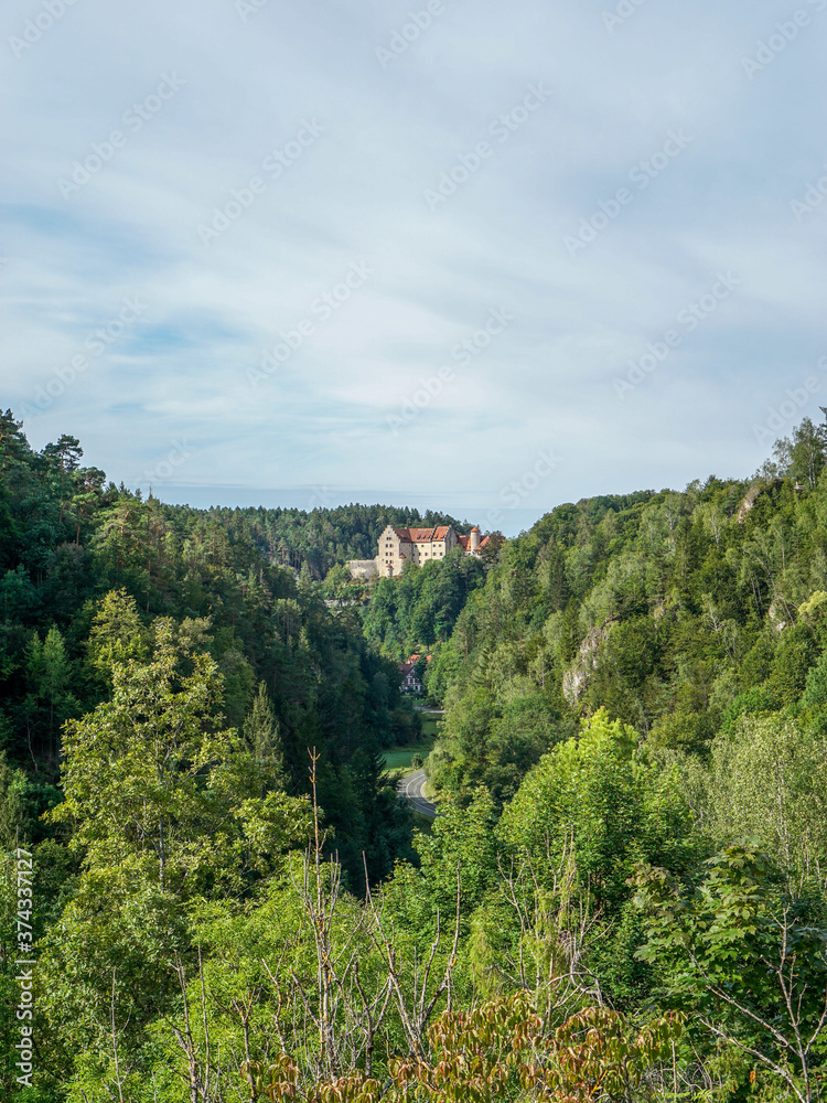 Straße am Bildrand führt zur Burg Rabenstein in der Fränkischen Schweiz bei Tag. Wald aus Nadelbäumen mit einer Burg als Motiv bei blauem Himmel und Tageslicht. Bayern, Deutschland.