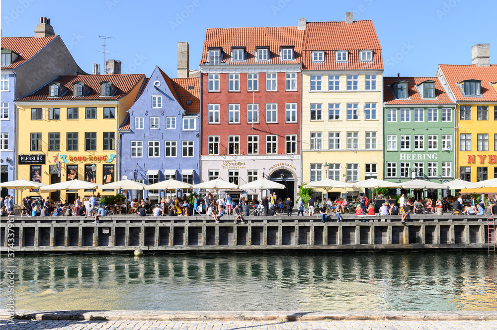 COPENHAGEN, DENMARK 29. June 2019: People relax on a promenade along Nyhavn in front of a row of colorful houses on a warm summer's day.