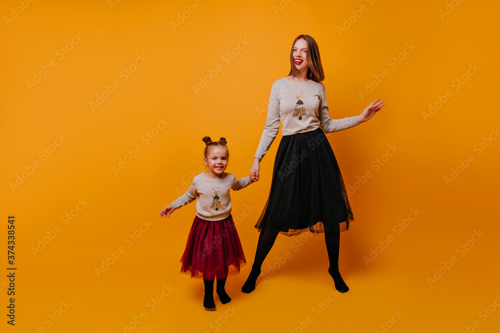 Young mother with a little girl of 4-6 years old is having fun and dancing in a New Year's clothes on an isolated orange background. Photo of happy mothers and daughters in full growth.