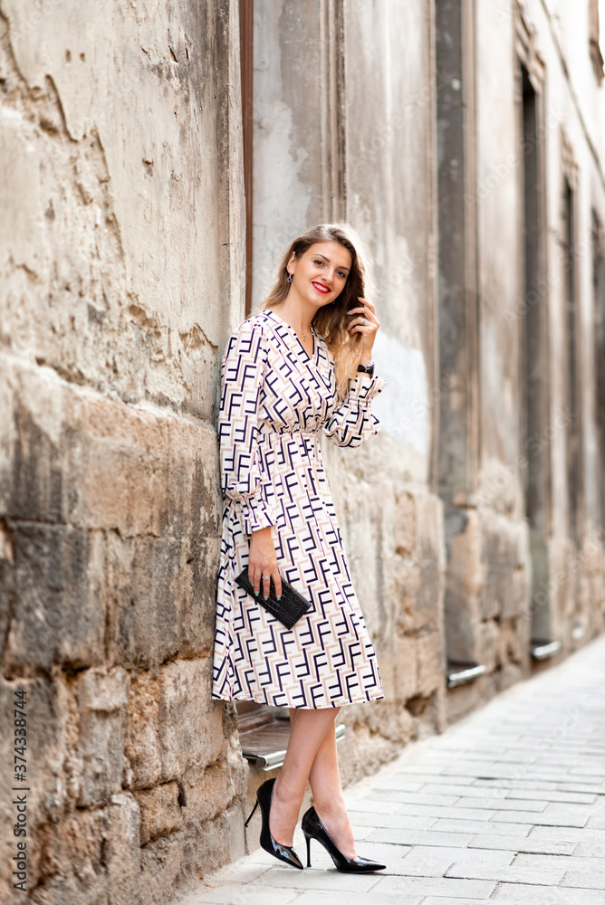A young beautiful woman with a small handbag in her hands stands near the wall of the old city.