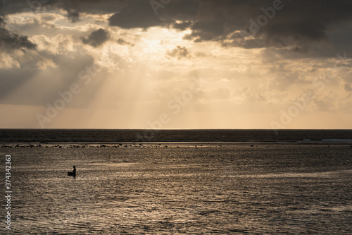 Ray of light above the shore broke through the black clouds. The sea is golden in color, and the atmosphere is quiet and serene. A sailor is looking for fish. © Pande