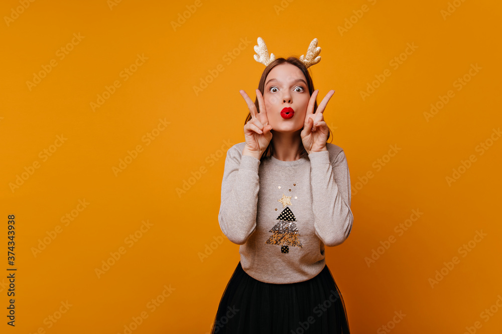 An enthusiastic, funny, young, perky girl 25 years old smiles into the camera and shows a peace sign with two hands near her face. Photo in studio on an orange background.