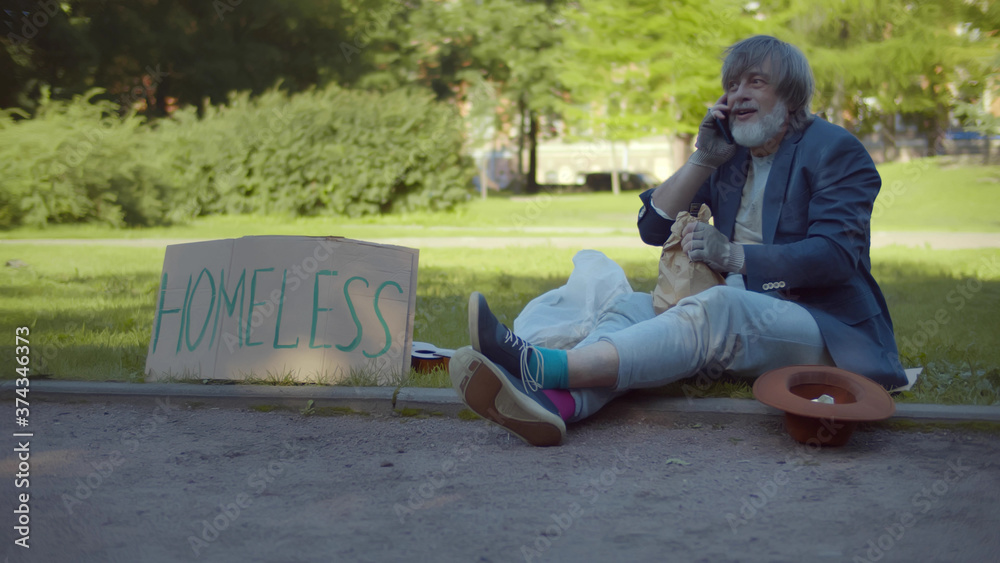 Homeless old man sitting on ground in park and talking on smartphone