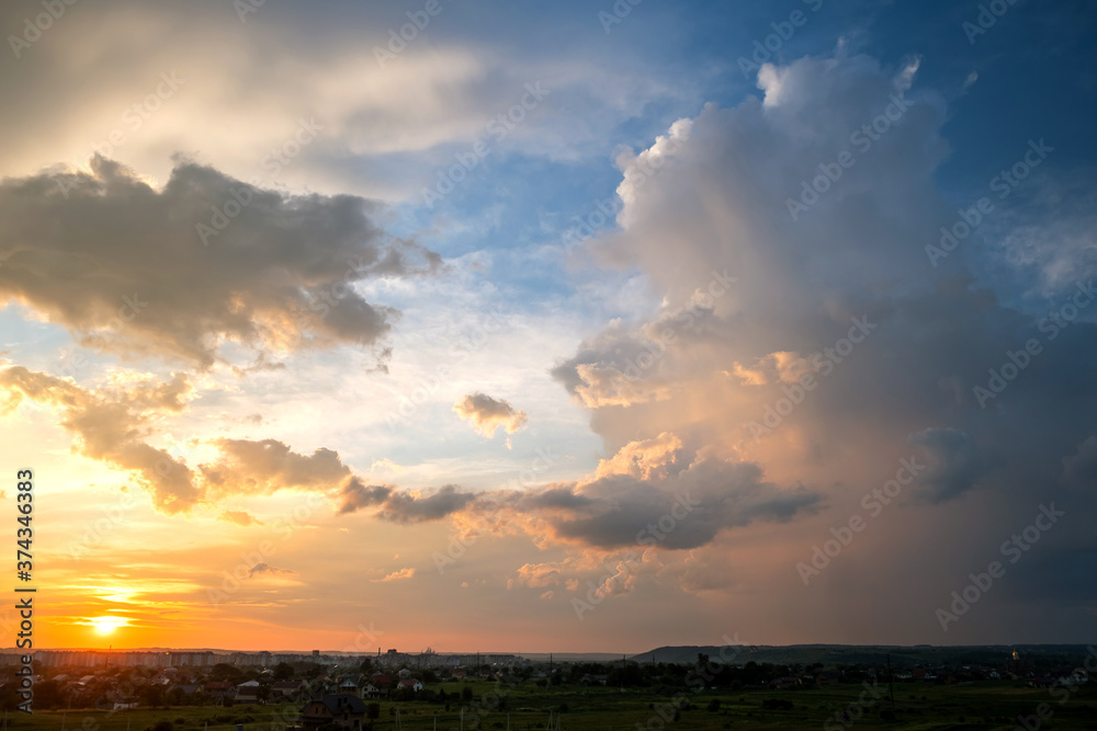 Dramatic sunset over rural area with stormy puffy clouds lit by orange setting sun and blue sky.