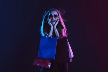 Astonished, shocked with shopping bags, cheerful. Portrait of young woman in neon light on dark backgound. The human emotions, black friday, cyber monday, purchases, sales, finance concept.